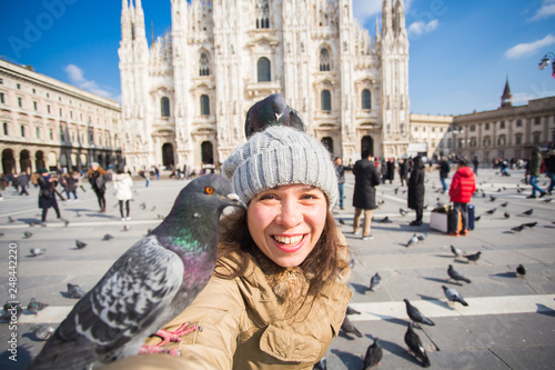 Winter travel, vacations and birds concept - Young happy woman tourist with funny pigeons making selfie photo in front of the famous Duomo cathedral in Milan.