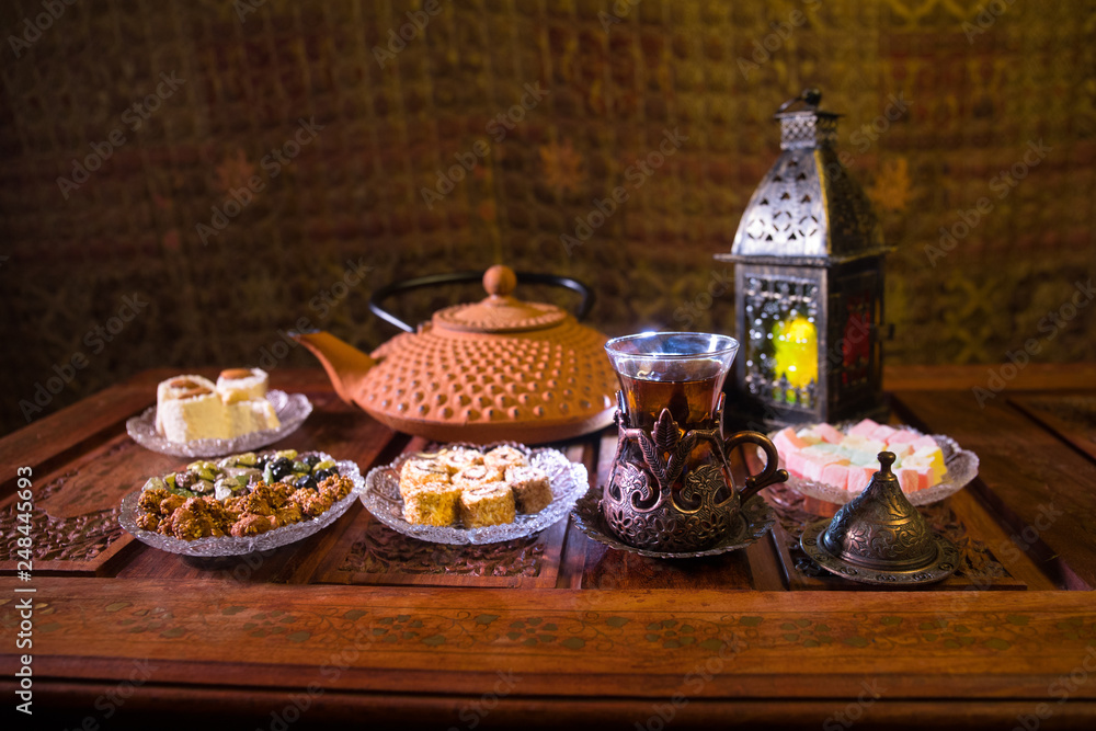 Arabian tea in glass with eastern snacks on vintage wooden surface. Eastern tea concept. Low light lounge interior with carpet. Empty space.