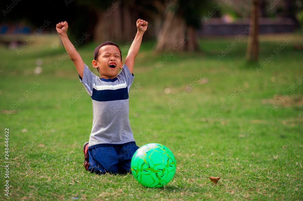 Obraz premium lifestyle portrait at grass city park of 5 years old Asian kid playing football happy and excited raising arms celebrating scoring goal in child sport practice education