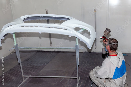A male worker paints with a spray gun a part of the car body in white after being damaged at an accident. Bamper from the vehicle during the repair in the workshop.
