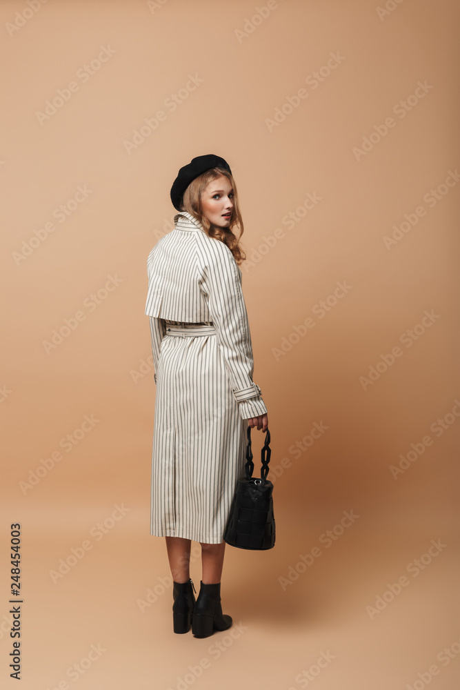 Young attractive woman in striped coat and black hat holding bag in hand thoughtfully looking in camera from back over beige background isolated Young attractive woman in striped coat and black hat holding bag in hand thoughtfully looking in camera from back over beige background isolated