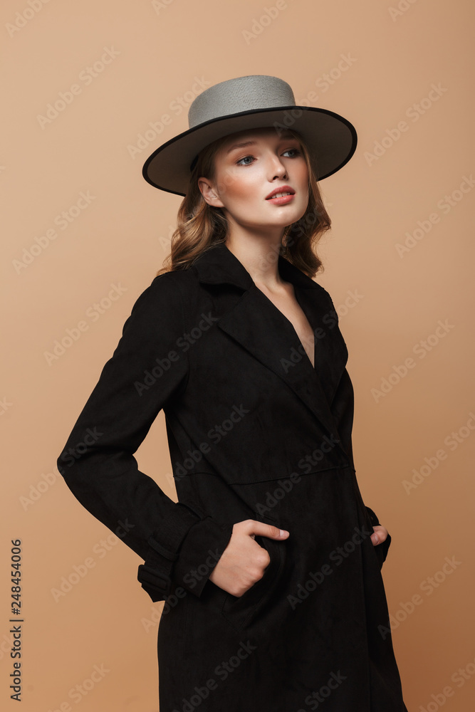 Fototapeta premium Young gorgeous woman with wavy hair in black coat and gray hat dreamily looking aside standing over beige background