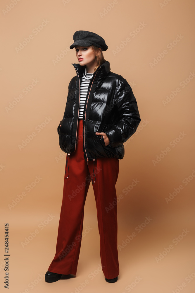 Young serious woman with wavy hair in cap,wide pants and black down jacket thoughtfully looking in camera standing over beige background Young serious woman with wavy hair in cap,wide pants and black down jacket thoughtfully looking in camera standing over beige background