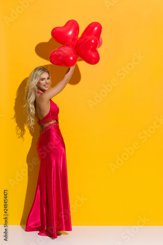 Beautiful Woman In Elegant Red Dress Is Holding Heart Shaped Balloons Over Her Head And Laughing