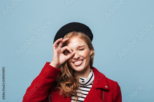 Close up beautiful smiling girl with wavy hair in black beret and red jacket playfully showing tongue while looking in camera over blue background