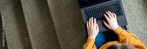 Tableau sur toile Young female college student sitting on stairs at school, writing essay on her laptop