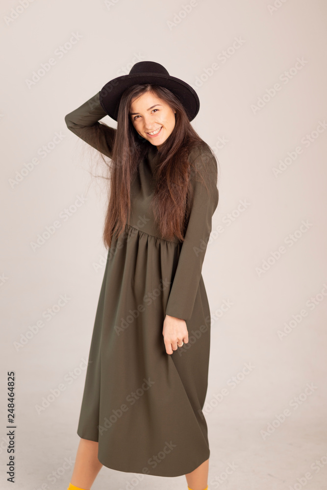 Young sensual caucasian woman in dark green (olive) dress holding fedora hat isolated on white-grey background with beautifull smile