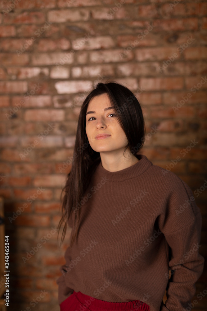 young, thin and attractive girl student dressed in casual clothes, red pants and a brown jacket, poses for a photo opposite the stone wall in the photo studio young, thin and attractive girl student dressed in casual clothes, red pants and a brown jacket, poses for a photo opposite the stone wall in the photo studio
