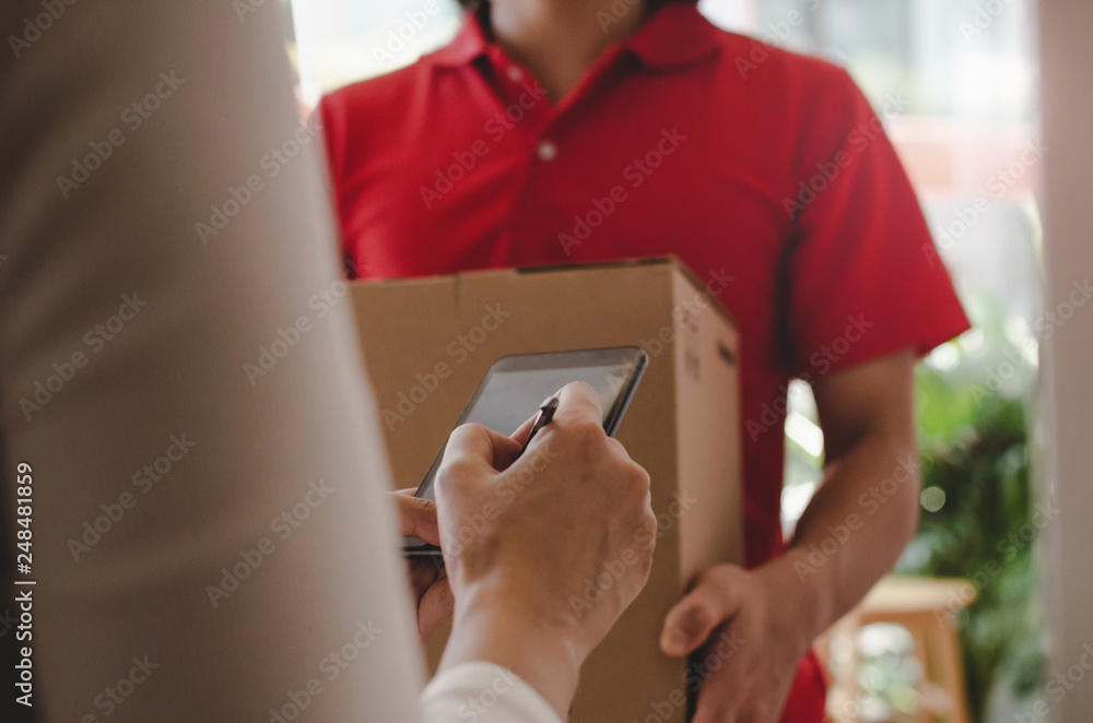 home delivery service man in red uniform and young woman customer appending signature in digital mobile phone receiving parcel post box from courier at home, express delivery, online shopping concept