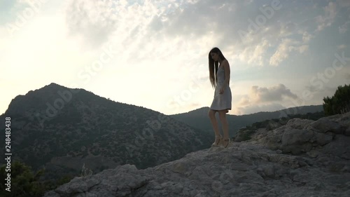 slim woman with long loose flowing hair in sundress walks on rock crest against bright sunlight from behind dark hills photo