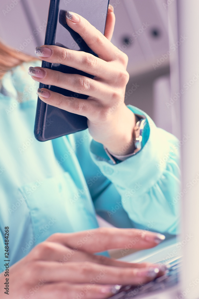 Young businesswoman having business call in office. Woman holds a smartphone in her hand and looks at the screen with her second hand typing on a laptop.