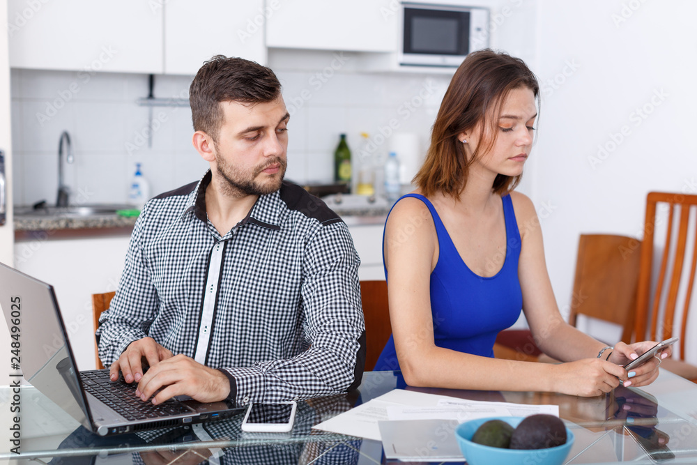 Fototapeta premium Young man working at laptop and upset girl using phone at home