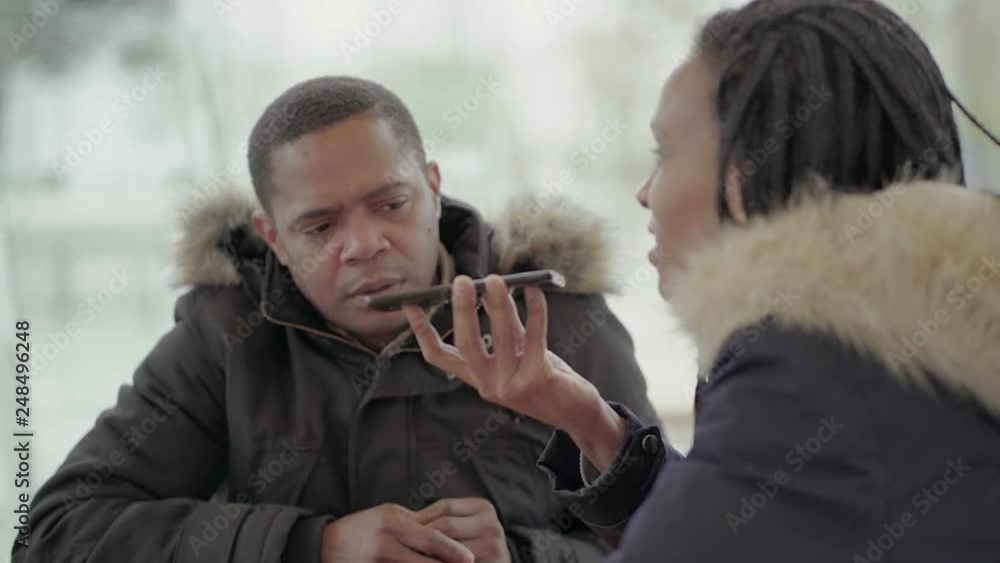 Afro-American young girl with braids and Afro-American middle-aged man in jacket with fur hood sitting outside, calling friend with speakerphone. Communication lifestyle, wireless technology concept Afro-American young girl with braids and Afro-American middle-aged man in jacket with fur hood sitting outside, calling friend with speakerphone. Communication lifestyle, wireless technology concept