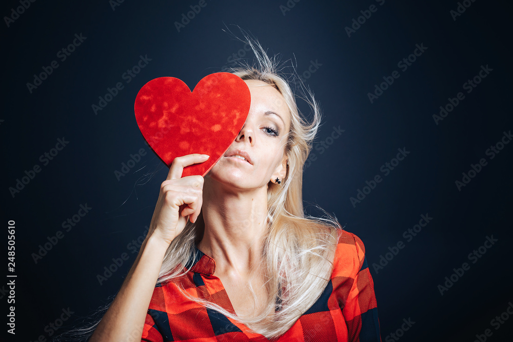 A girl on a black background with a Valentine in the form of a heart covers one eye.