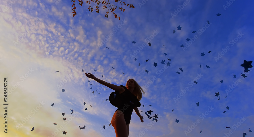 A young girl stands outdoors at sunset. She happily throws blue paper stars in the air to express her emotions and excitement.