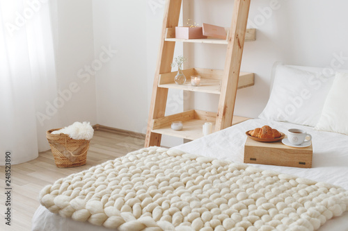 Stylish cozy scandinavian bedroom interior: bed with white linen, light beige thick yarn knitted woolen merino chunky blanket, wooden rack, tray with a croissant and a cup of tea, white wall. 
