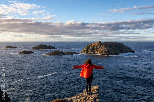 Woman in red jacket is standing at the edge of a cliff with open arms and enjoying the beautiful ocean scenery. Taken in Crow Head, North Twillingate Island, Newfoundland and Labrador, Canada.