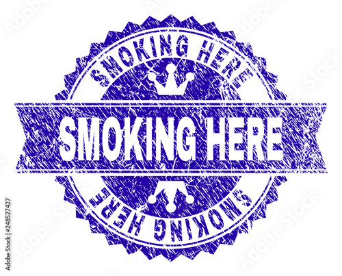 SMOKING HERE rosette stamp seal watermark with distress texture. Designed with round rosette, ribbon and small crowns. Blue vector rubber watermark of SMOKING HERE label with dirty texture.