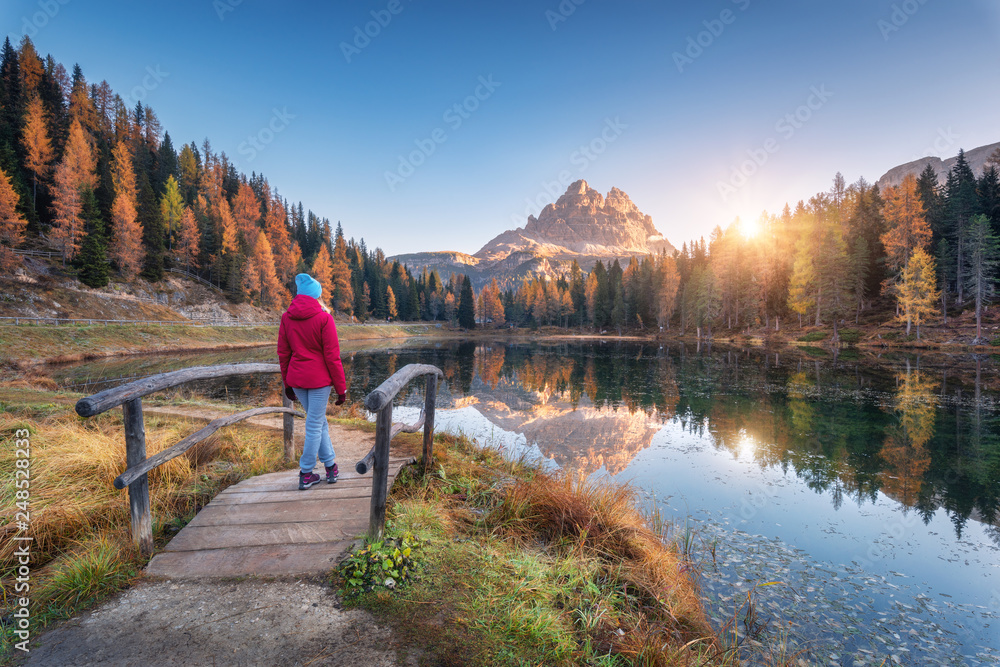 Young wooman on the small wooden bridge against lake, autumn forest, mountain peak, blue sky with sunlight at sunrise. Landscape with girl, reflection in water, green grass, rocks in Dolomites, Italy