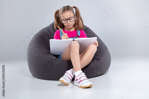 Young girl in glasses doing homework on a gray bean bag