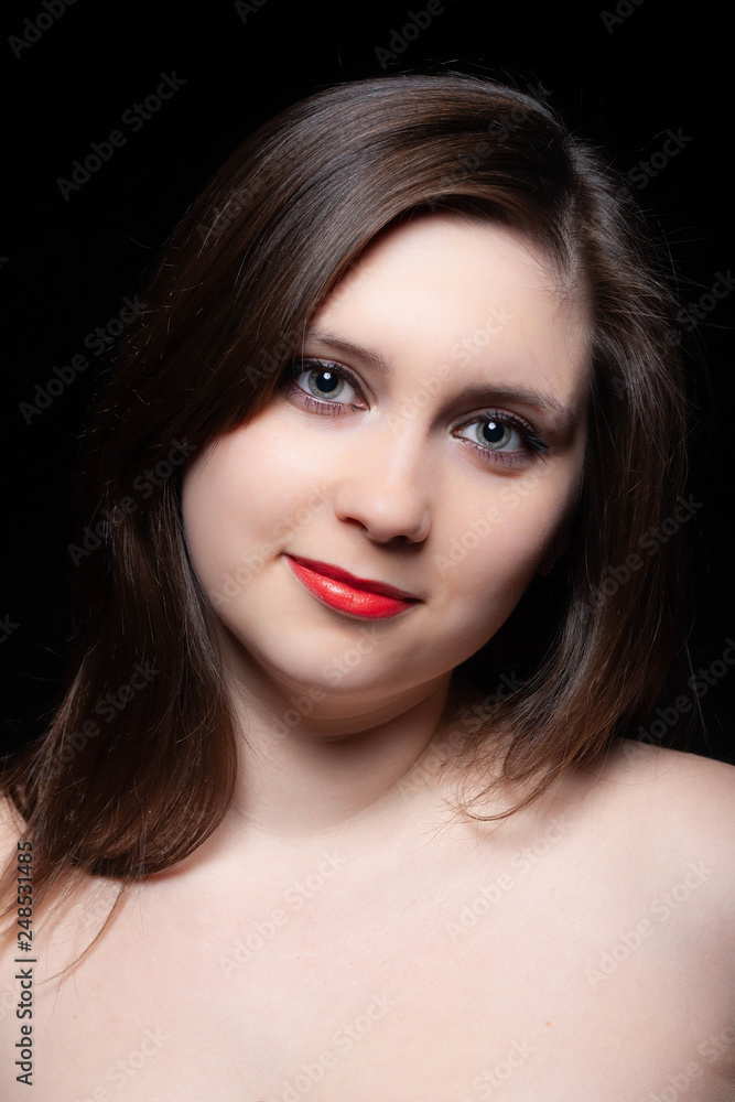 Beautiful gray-eyed brown-haired woman with red lips and bared shoulders against black background. Closeup studio portrait