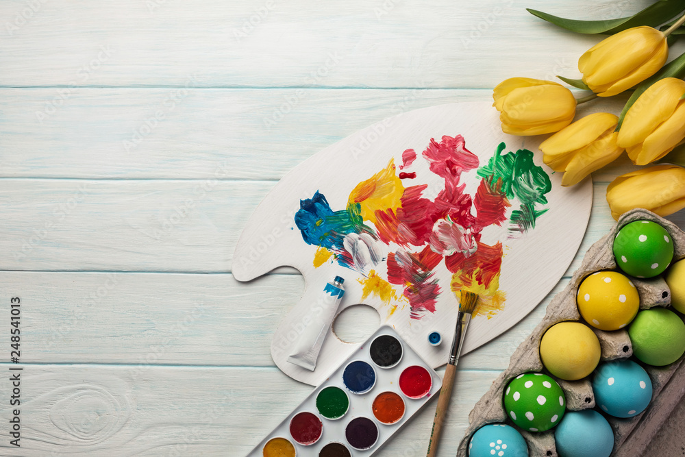 Colorful easter eggs, paint palette, brushes and yellow tulips on white wooden table. Top view with copy space. Colorful easter eggs, paint palette, brushes and yellow tulips on white wooden table. Top view with copy space.