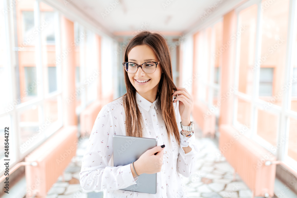 Cute young female teacher holding tablet and touching her brown hair while looking away. University hallway interior. Cute young female teacher holding tablet and touching her brown hair while looking away. University hallway interior.