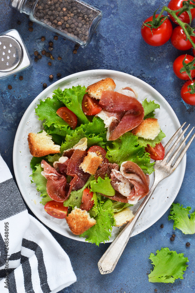 Salad with prosciutto, cherry tomatoes, bread crisps on the kitchen table. View from above.