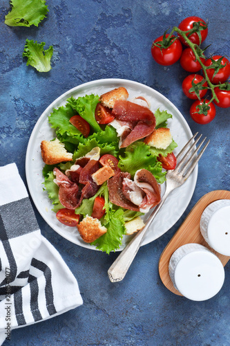 Salad with prosciutto, cherry tomatoes, bread crisps on the kitchen table. View from above.