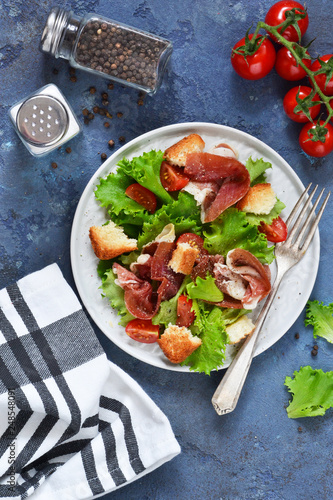 Salad with prosciutto, cherry tomatoes, bread crisps on the kitchen table. View from above.