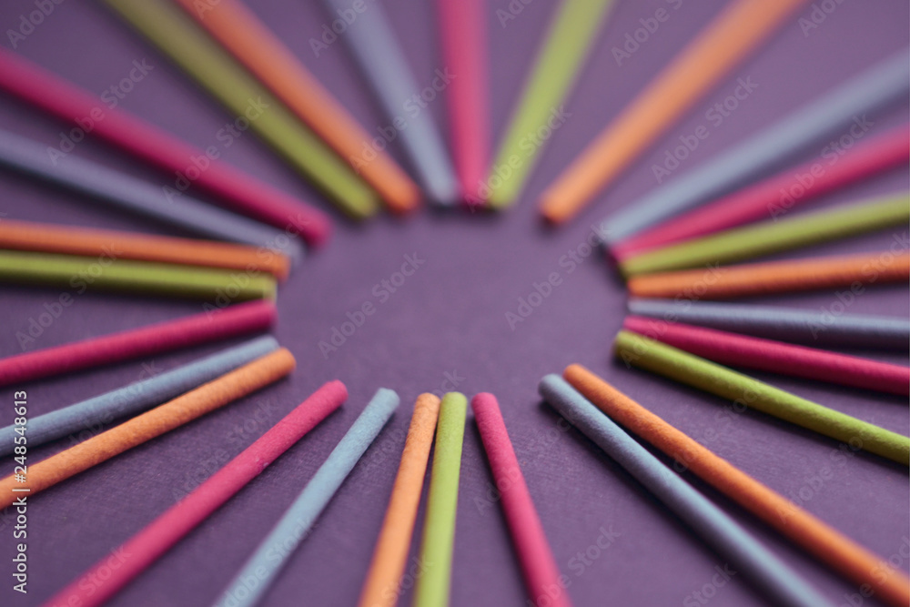Multicolored Indian incense sticks lie in the shape of sun and rays on a dark purple background, with empty circle
