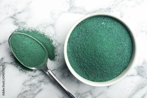 Bowl and spoon of spirulina algae powder on marble background, top view