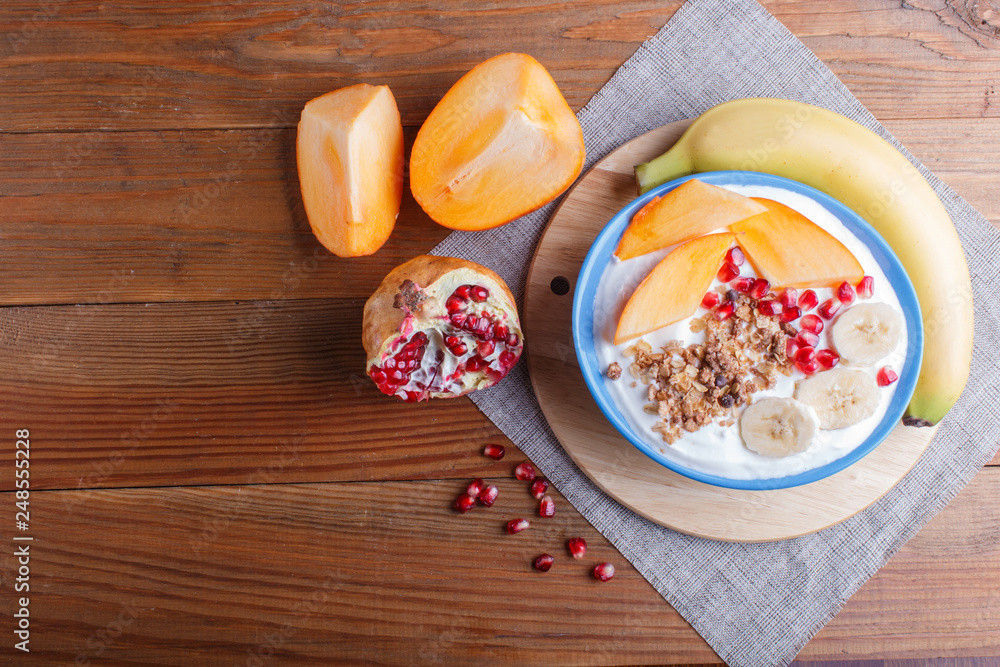 A blue plate with greek yogurt, granola, persimmon, banana, pomegranate on brown wooden background.