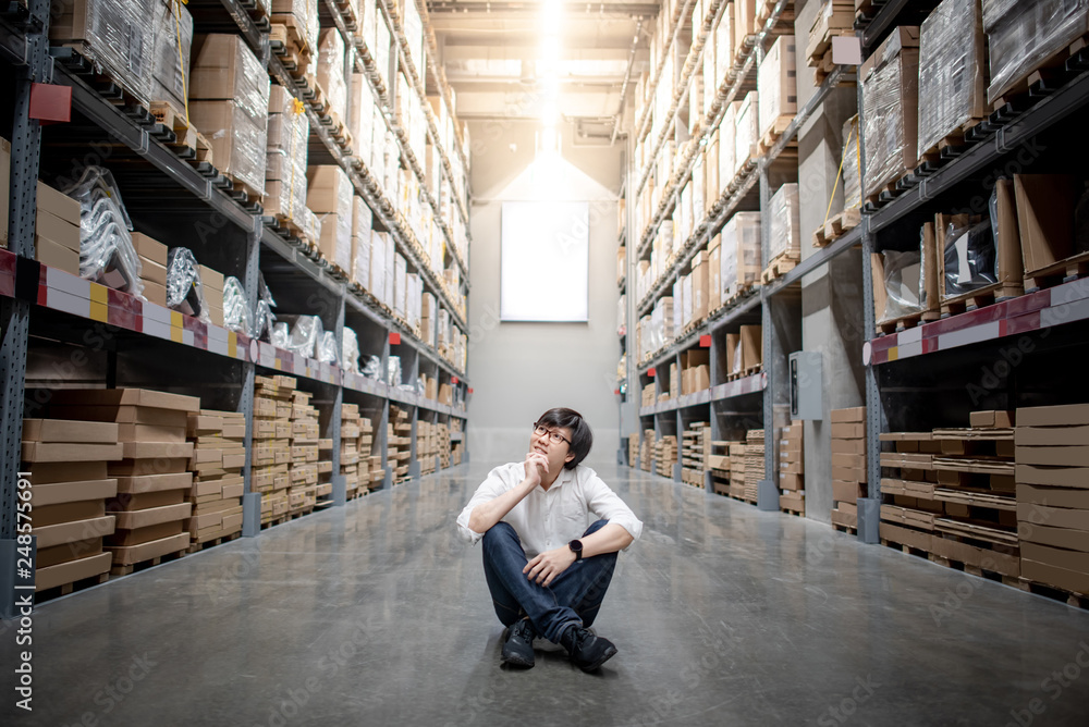 Fototapeta premium Asian shopper man sitting between cardboard box shelves aisle in warehouse choosing what to buy. Shopping lifestyle in department store. Buying or purchasing factory goods. Inventory industry concept