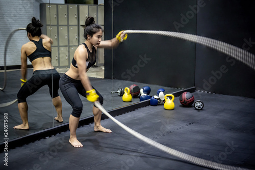 Young Asian girl doing exercise with battle rope in fitness gym. Physical workout with muscular build activity. Female bodybuilding concept