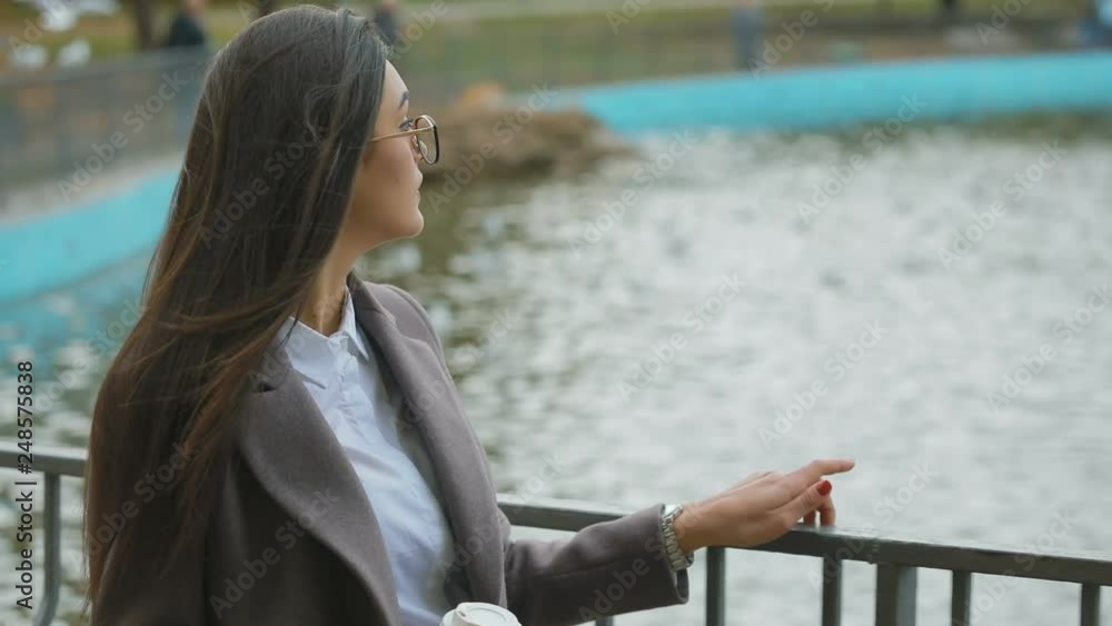 slow motion young brunette woman in coat with paper cup walks and holds on to handrail against park pond slow motion young brunette woman in coat with paper cup walks and holds on to handrail against park pond