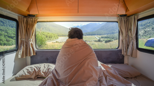 Young Asian man traveler staying in the blanket looking at mountain scenery through the window in camper van in the morning. Road trip in summer of South Island, New Zealand.