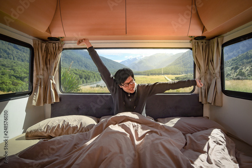 Lazy Asian man traveler stretching arms while staying in the blanket in camper van in the morning with mountain scenery in the background. Road trip in summer of South Island, New Zealand.