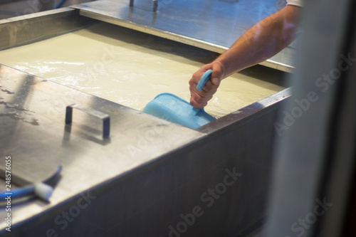 Technology of making cheese from milk on factory, shot through glass of aceptic room, men's hand mixes and cooks cheese with big turner in huge metal tub. Horizontal. Flecks on glass. photo