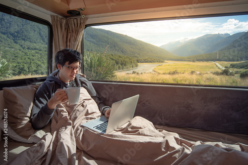 Young Asian man traveler drinking hot coffee while working with laptop computer on the bed in camper van with mountain scenic view through the window. Digital nomad or freelance on road trip concept © zephyr_p