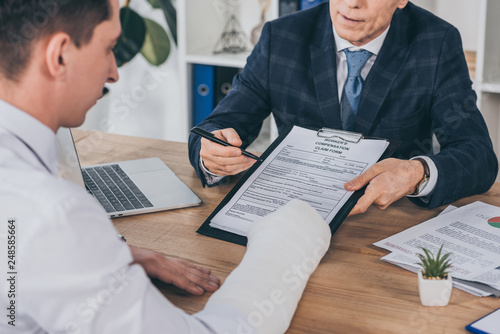businessman in blue jacket giving form for compensation claim to worker with broken arm at table in office, compensation concept photo