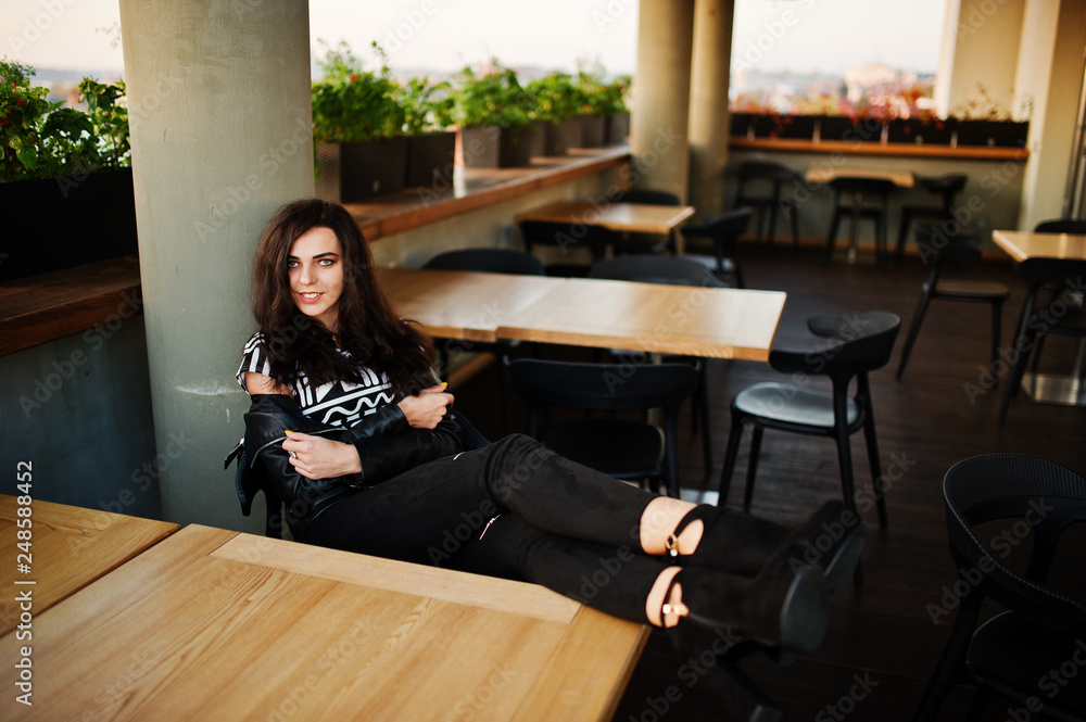 Young curly woman in leather jacket in a bar put her legs on table. Young curly woman in leather jacket in a bar put her legs on table.