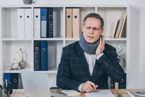 Cuadro en lienzo businessman in neck brace sitting at table, feeling pain and holding neck in off