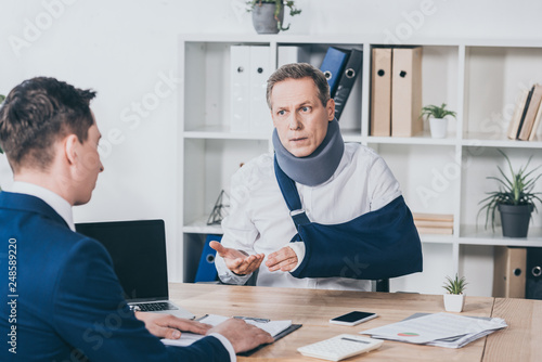 middle aged worker in neck brace with broken arm sitting at table and talking to businessman in blue jacket in office, compensation concept photo