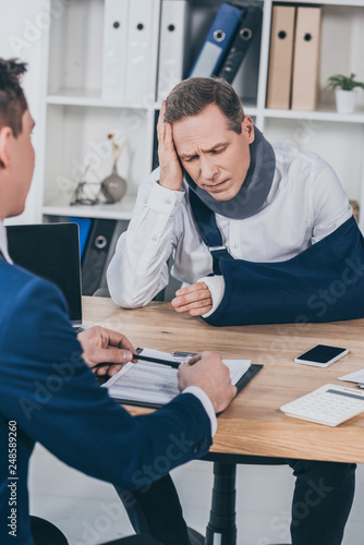 upset worker in neck brace with broken arm sitting at table opposite businessman in blue jacket in office, compensation concept © LIGHTFIELD STUDIOS