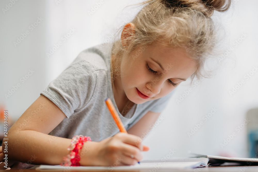 A seven-year-old girl sits at home at a table and writes in a notebook, completing a learning task or repeating lessons. A seven-year-old girl sits at home at a table and writes in a notebook, completing a learning task or repeating lessons.
