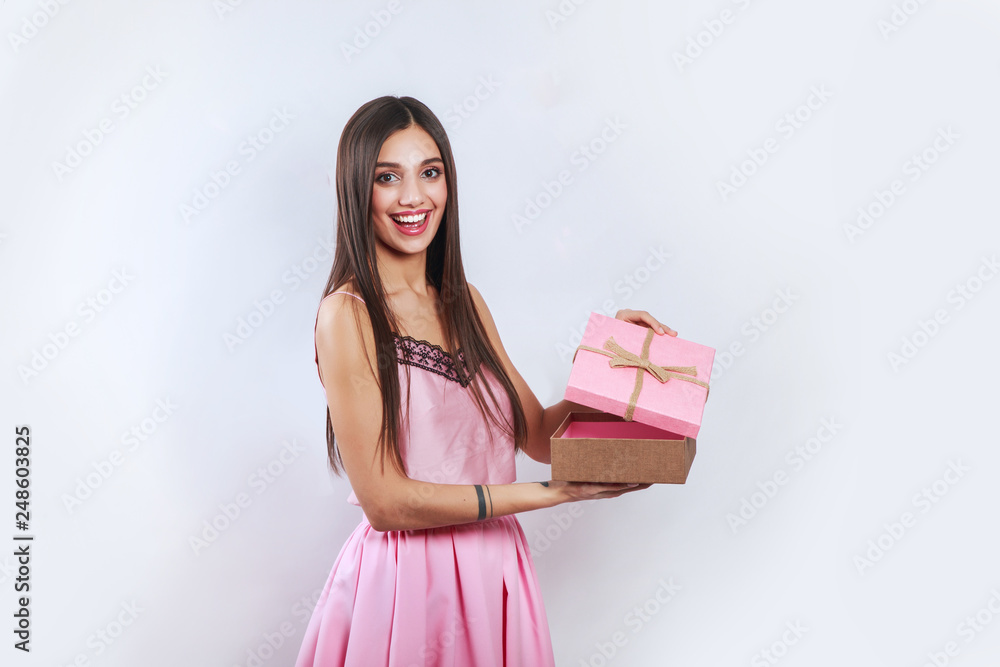 Surprised brunette woman holding pink gift and looking on camera over gray background Surprised brunette woman holding pink gift and looking on camera over gray background