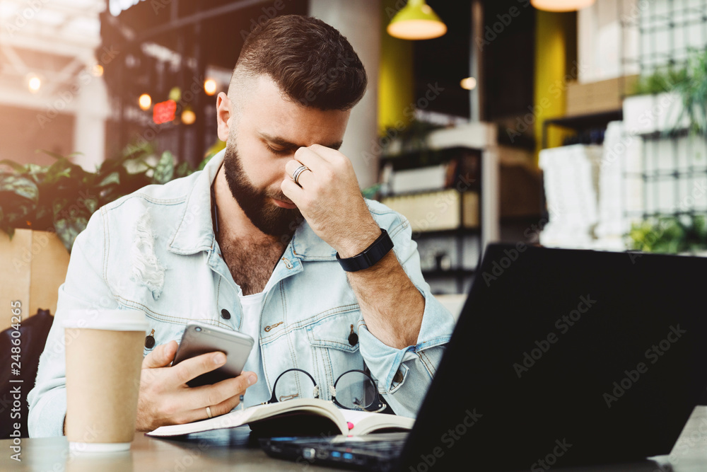Young bearded sad businessman is sitting at table, covering his face with his hand and holding smartphone in his hand. Man got bad news, problems. Stressed.On desk is laptop, notepad, cup of coffee. Young bearded sad businessman is sitting at table, covering his face with his hand and holding smartphone in his hand. Man got bad news, problems. Stressed.On desk is laptop, notepad, cup of coffee.