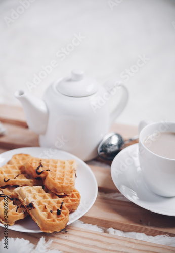 Cozy breakfast in bed, cup of coffee and heart shaped waffles on wooden tray on white and gray cozy blanket, the concept of home comfort, copy space