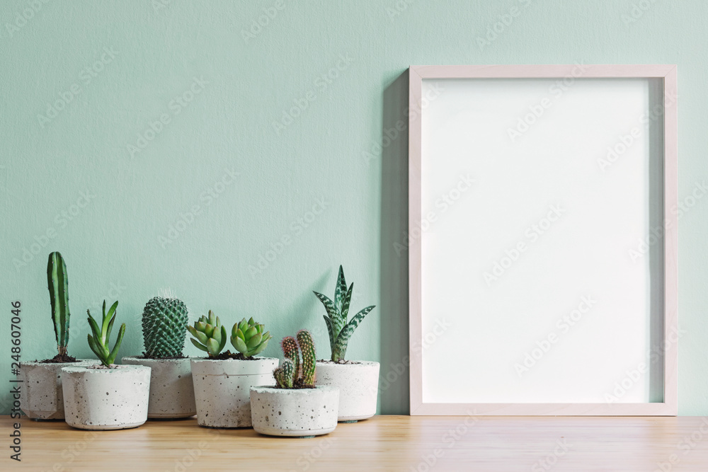 Minimalistic home interior with mock up photo frame on the brown wooden table with composition of cacti and succulents in stylish cement pots. Mint walls. Stylish concept of home garden.